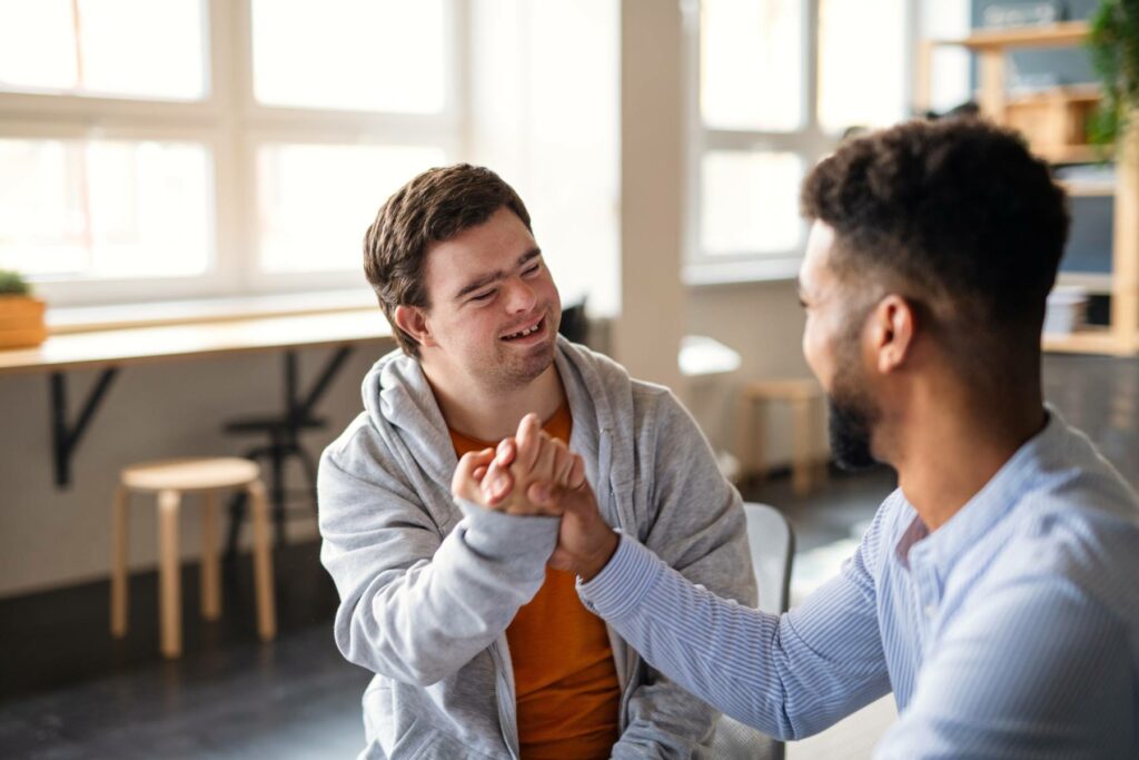 Young happy man with Down syndrome with his mentoring friend celebrating success indoors at school.